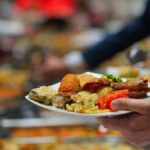 En tallrik full av buffémat, hållen av hand med buffébord i bakgrunden.