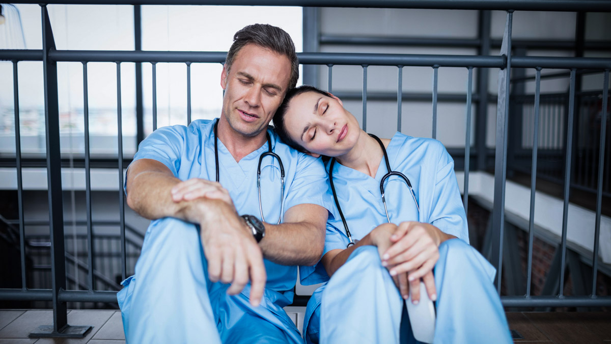 Två läkare sitter utanför en byggnad och vilar sig mot varandra.