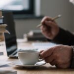 Man med vitt skägg sitter med penna och kaffekopp framför dator. En hög böcker står intill, på bordet.