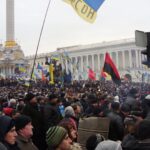 Folksamling på ett stort torg med vita byggnader i bakgrunden. Ukrainsk blågul flagga skymtas.