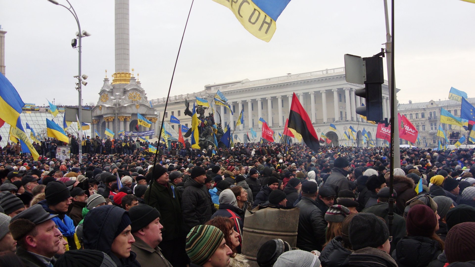 Folksamling på ett stort torg med vita byggnader i bakgrunden. Ukrainsk blågul flagga skymtas.