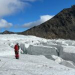 Två personer går på en glaciär med stora sprickor i. Berg utan snö och is syns i bakgrunden.