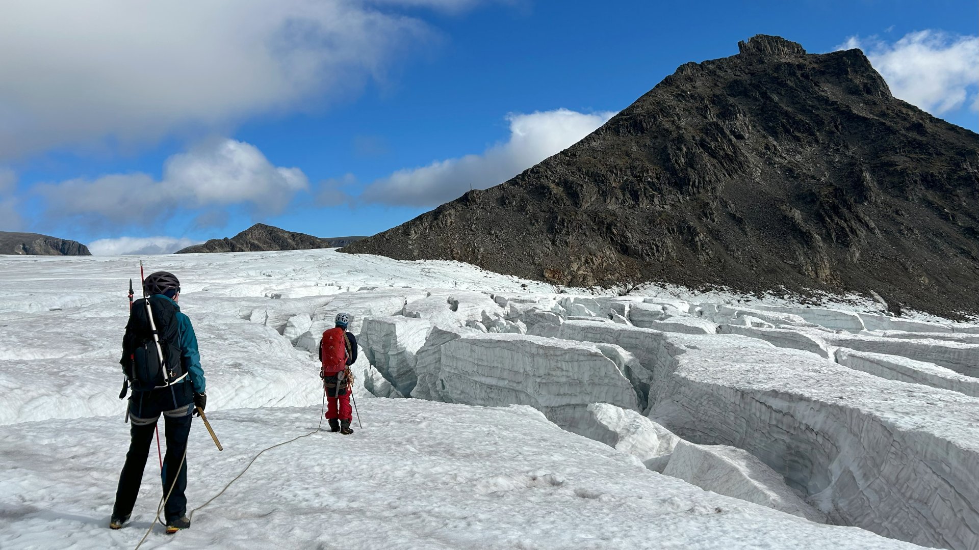 Två personer går på en glaciär med stora sprickor i. Berg utan snö och is syns i bakgrunden.