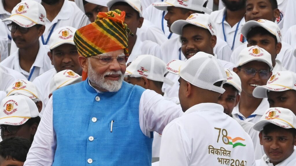 Premiärminister Narendra Modi i vit skjorta, blå väst och rödrandig huvudbonad hälsar på en vitklädd person i en folksamling.