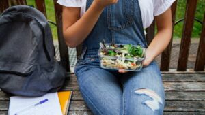 Ung person sitter på bänk med ryggsäck och skrivblock intill. Bär jeansdress och äter ut en matlåda med sallad och makaroner.