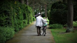 Äldre person i långbyxor och jacka leder cykel genom grön och lummig park.