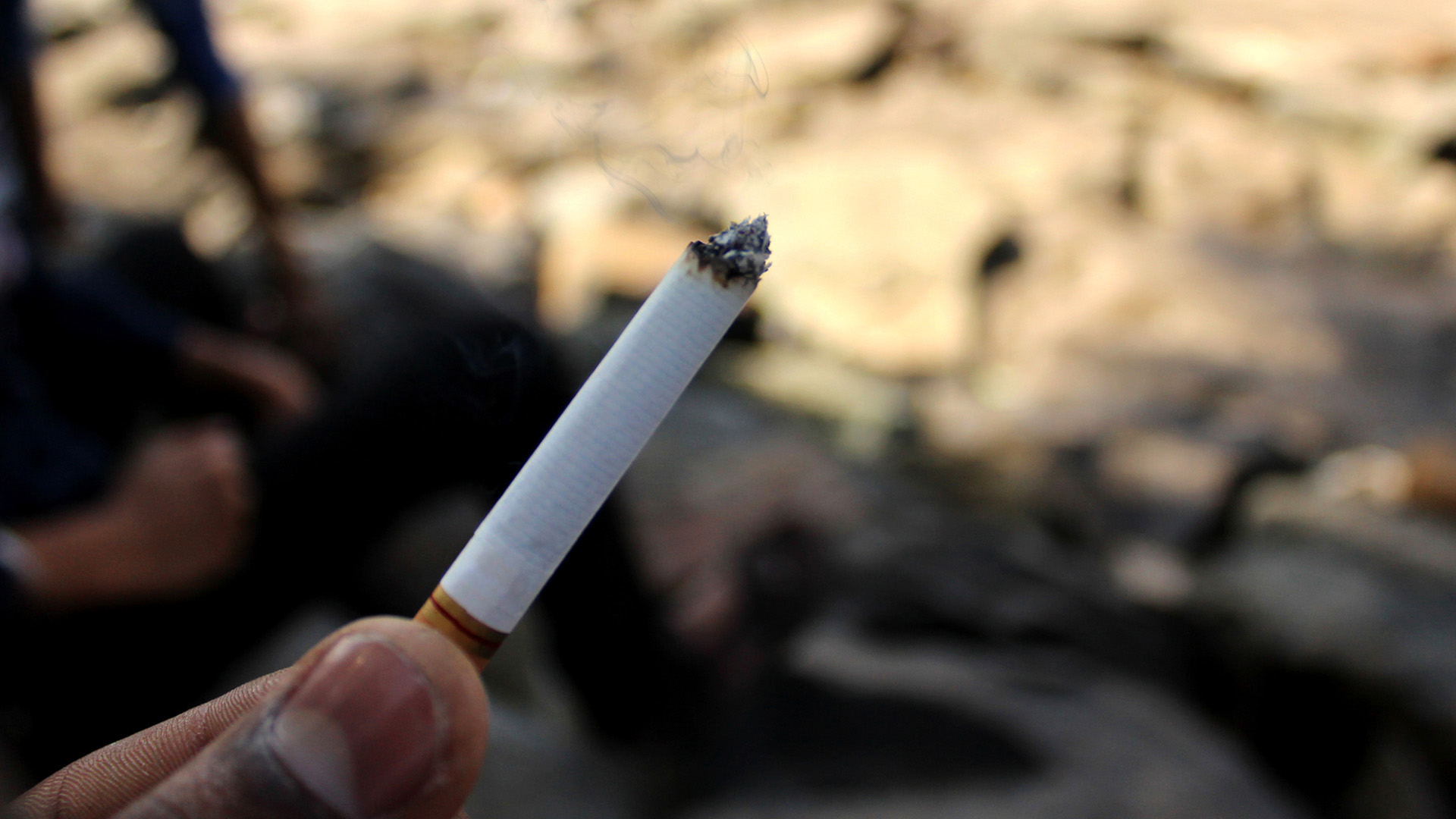 En person håller cigarett med rök mot suddig bakgrund.