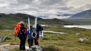 tre forskare med mätutrustning på fjället med en sjö i bakgrunden.