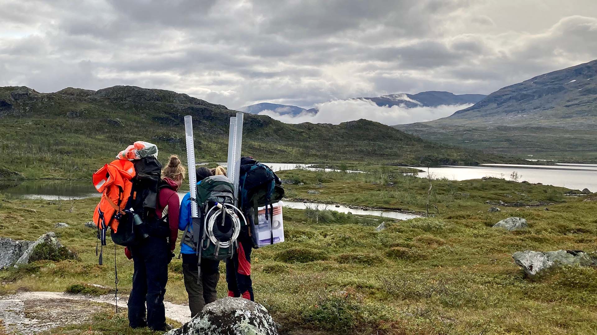 tre forskare med mätutrustning på fjället med en sjö i bakgrunden.