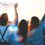 Ryggtavlor på tre unga tjejer som tittar ut över ett räcke i solnedgången.