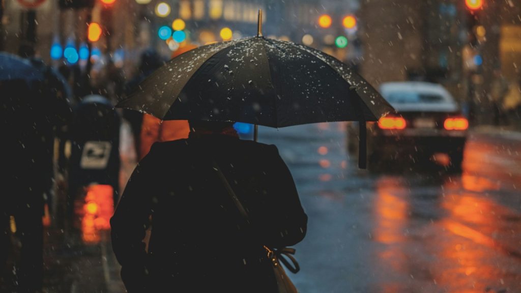 Person under paraply i stadsmiljö, snö faller, mörker och ljus och från billyktor och gatlampor.