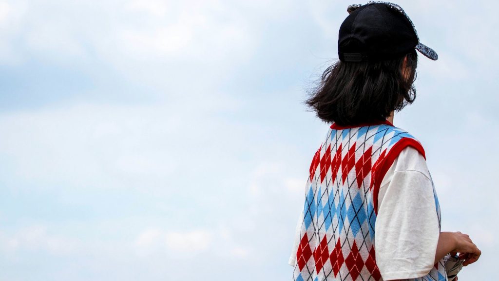 Ung mörkhårig person i keps, vit skjorta och färgglad väst står med ryggen mot kameran, mot himmel.