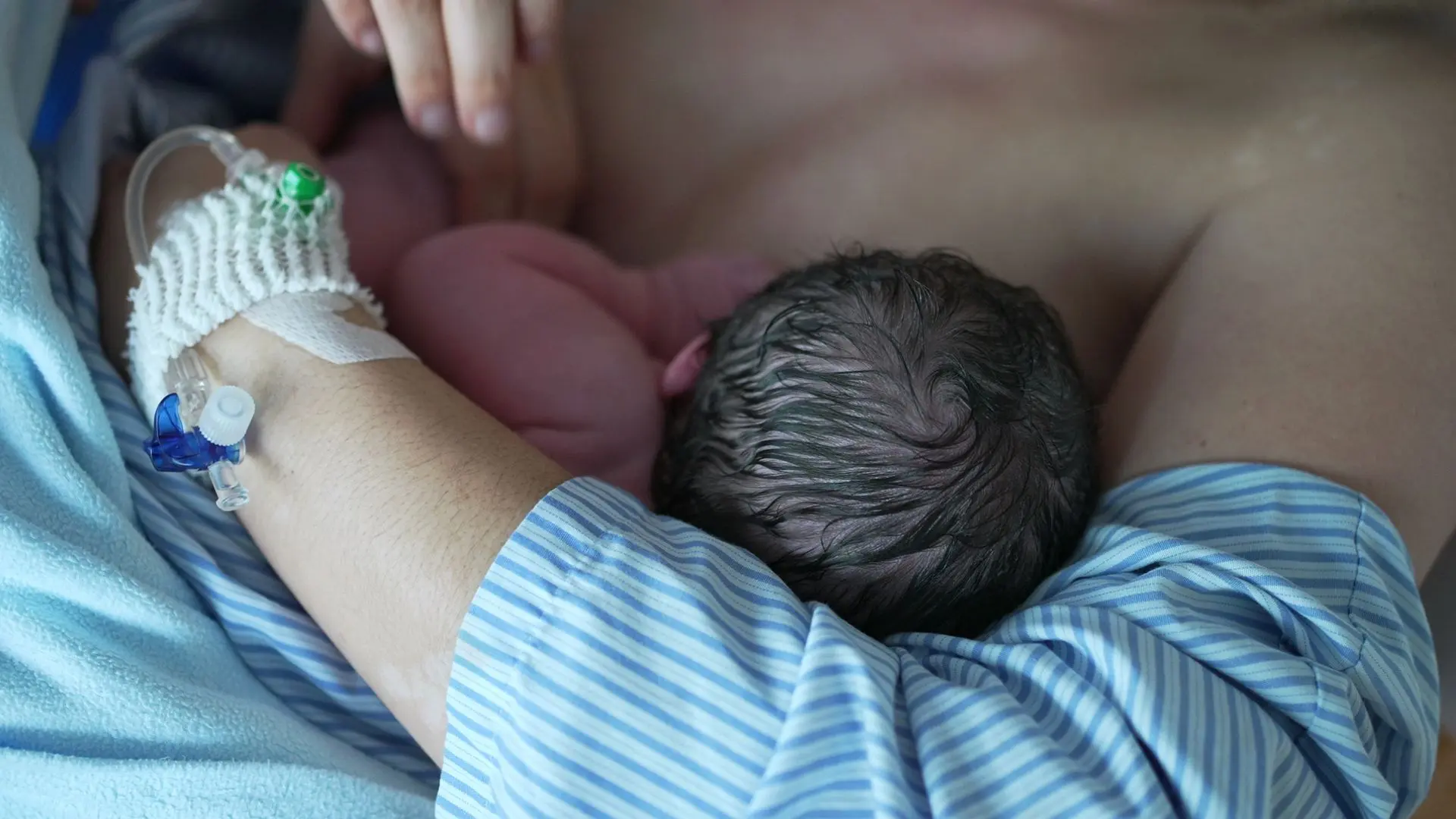 En nyfödd ligger vid mammans bröst, sjukhusmiljö med infart i den vuxnas hand.