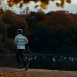 Ung man springer i t-shirt och byxor vid sjö, höstlöv på träd invid.