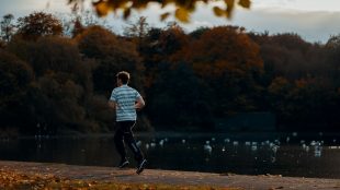 Ung man springer i t-shirt och byxor vid sjö, höstlöv på träd invid.