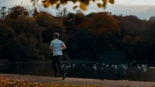 Ung man springer i t-shirt och byxor vid sjö, höstlöv på träd invid.