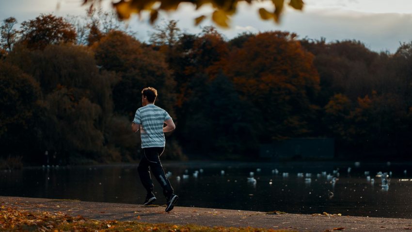 Ung man springer i t-shirt och byxor vid sjö, höstlöv på träd invid.