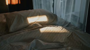 Morgonljus landar fläckvis på en säng med en vit huvudkudde och vitt lakan, gardin bakom.