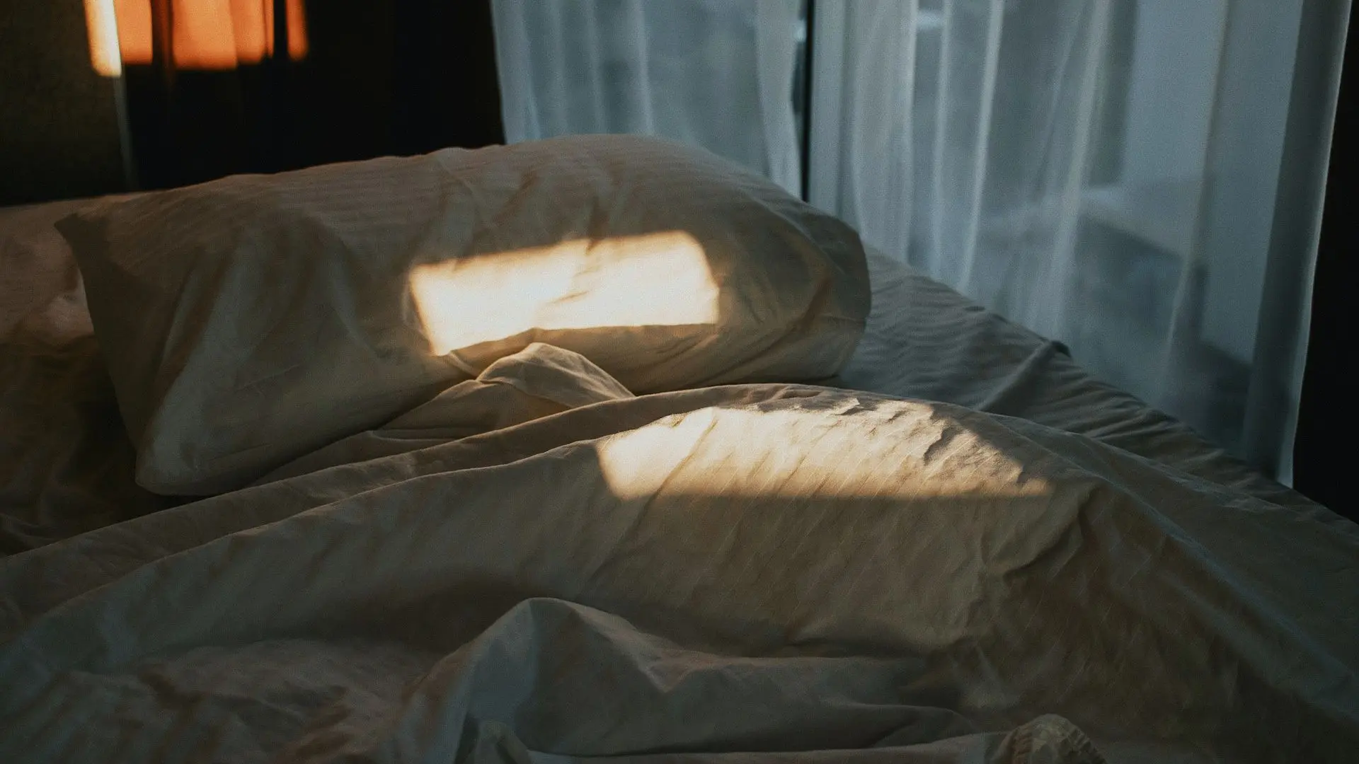 Morgonljus landar fläckvis på en säng med en vit huvudkudde och vitt lakan, gardin bakom.