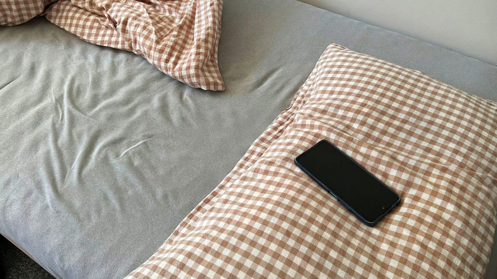 Madrass med rutig kudde och rutigt täcke. Svart mobiltelefon ligger på kudden.