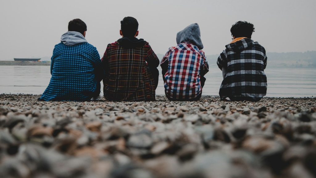 Fyra ungdomar i varma kläder sitter på stenig strand, vända mot vattnet.