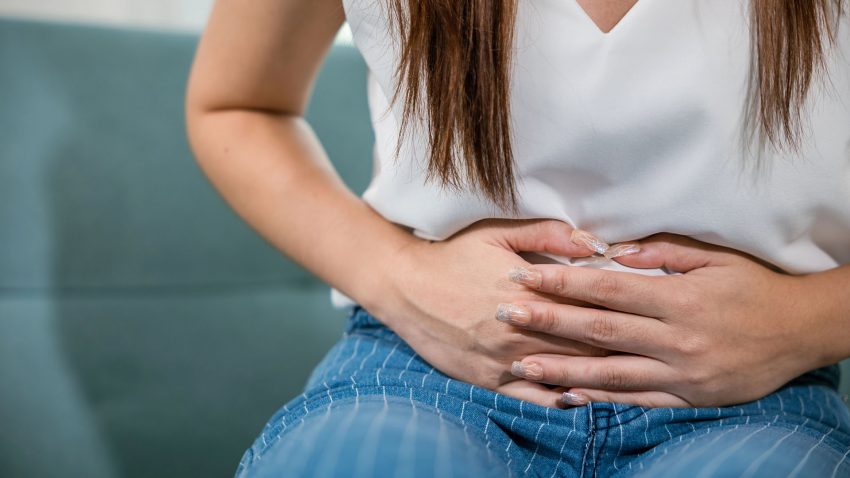 Ung kvinna i vit tröja håller händerna över magen.