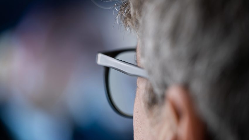 Man med grått hår och glasögon tittar mot en suddig skärm.