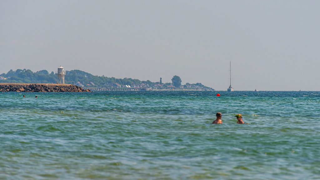 Två personer badar i grönblått vatten sommartid, stadssiluett i bakgrunden.