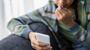 Barn med rutig tröja sitter i soffa och tittar på mobiltelefon i handen.