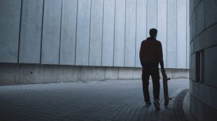 Man i svart siluett står med skateboard i handen i betongtunnel.