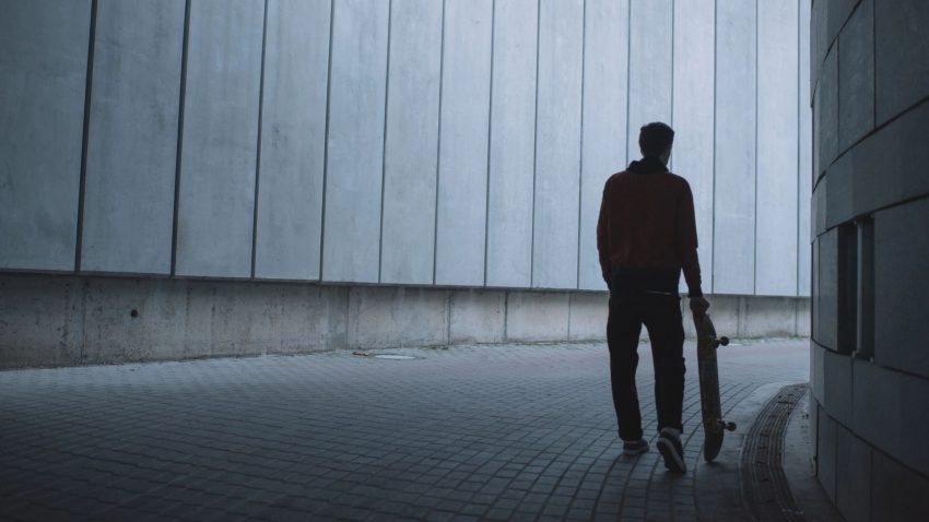 Man i svart siluett står med skateboard i handen i betongtunnel.