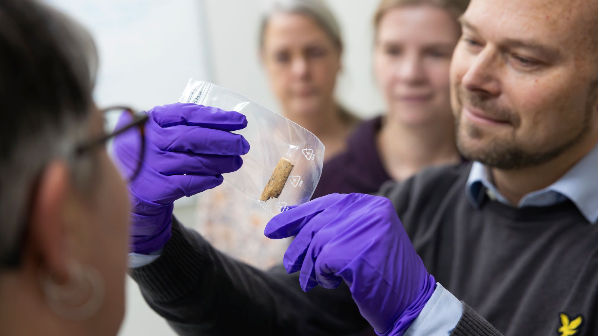 En person med mörklila handskar håller upp en genomskinlig påse med en brunt bit inuti. Tre andra personer tittar på biten.