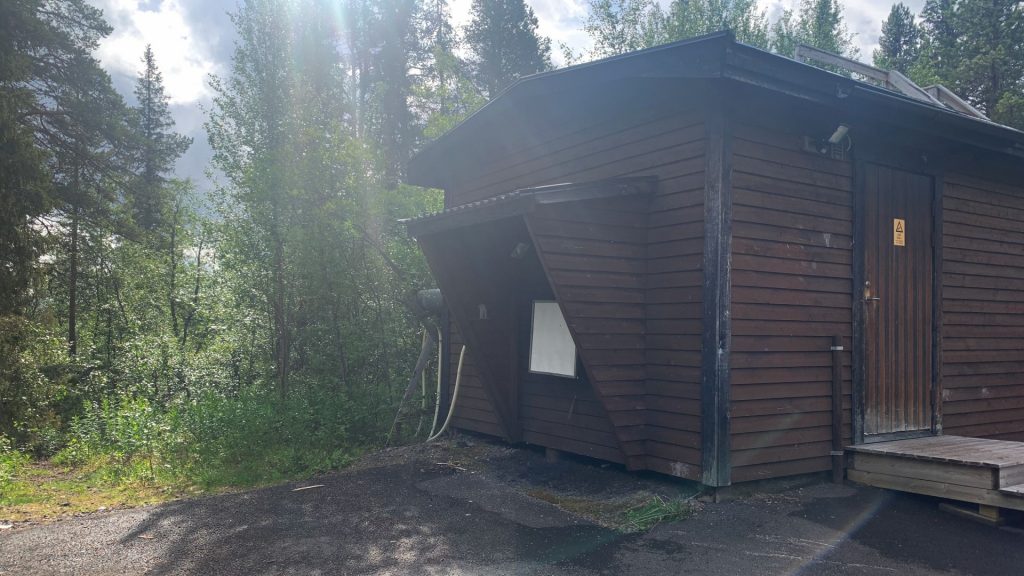 Brun stuga utan fönster, i skog. På en vägg sitter en vit tavla, ett filter.