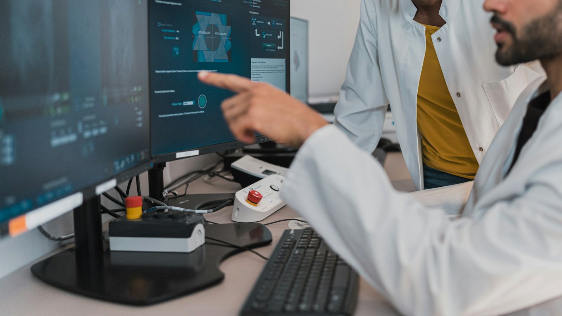 En person i vit rock, vid skrivbord, pekar mot en datorskarm. Annan person i vit rock i bakgrunden.