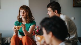 Tre unga personer sitter i hemmiljö och pratar. Ung kvinna med långt hår och tekopp säger något.