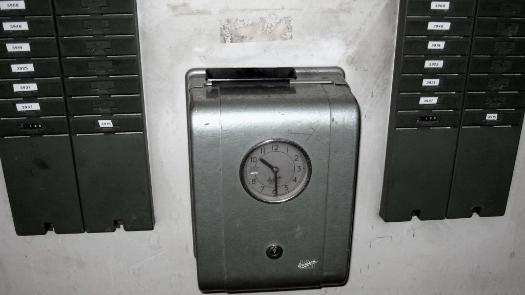 Stämpelklocka på vägg, svartvit bild. På väggen finns fack för stämpelkort.