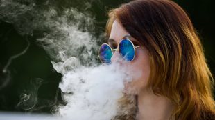 Tjej med brunt hår och blåtonade runda glasögon röker en e-cigarett.