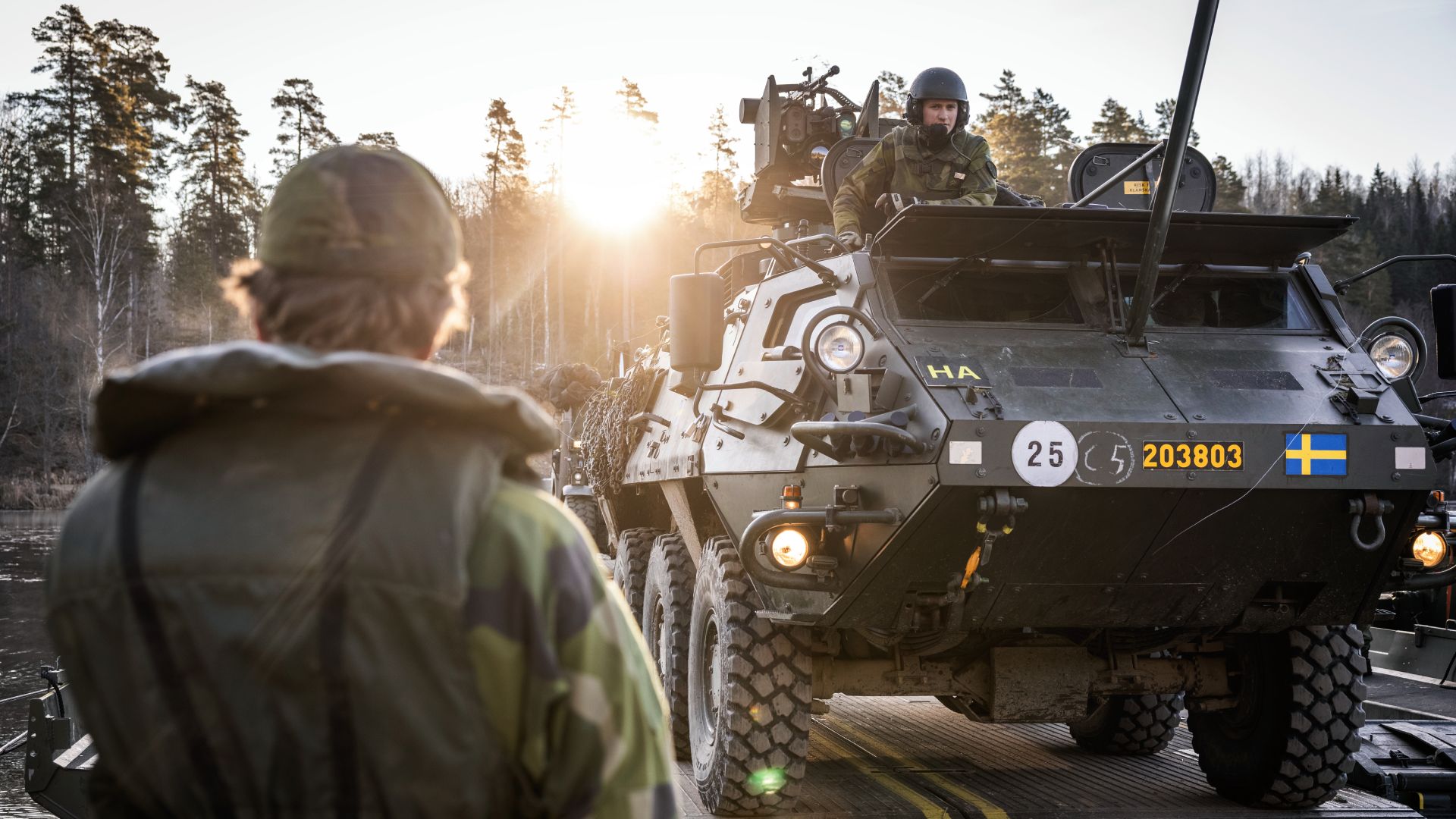 En ung person i militärkläder står med ryggen mot kameran. Bakom syns en stridsvagn med ung person i militärkläder högst upp.