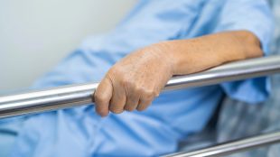 Äldre kvinna håller i grind till sjukhussäng.