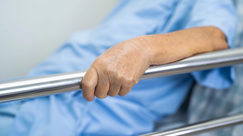 Äldre kvinna håller i grind till sjukhussäng.