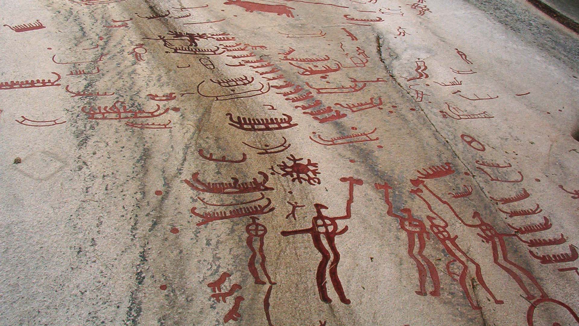 Ristningar med manliga figurer, båtar och djur på en stenhäll.