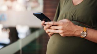 Gravid kvinna står ute på en balkong med mobil i handen.