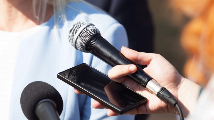 Reportrar med mikrofoner och inspelningsutrustning intervjuar en kvinna.