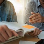 Två personer sitter vid bord och tittar i en bok med post it-lapp på.