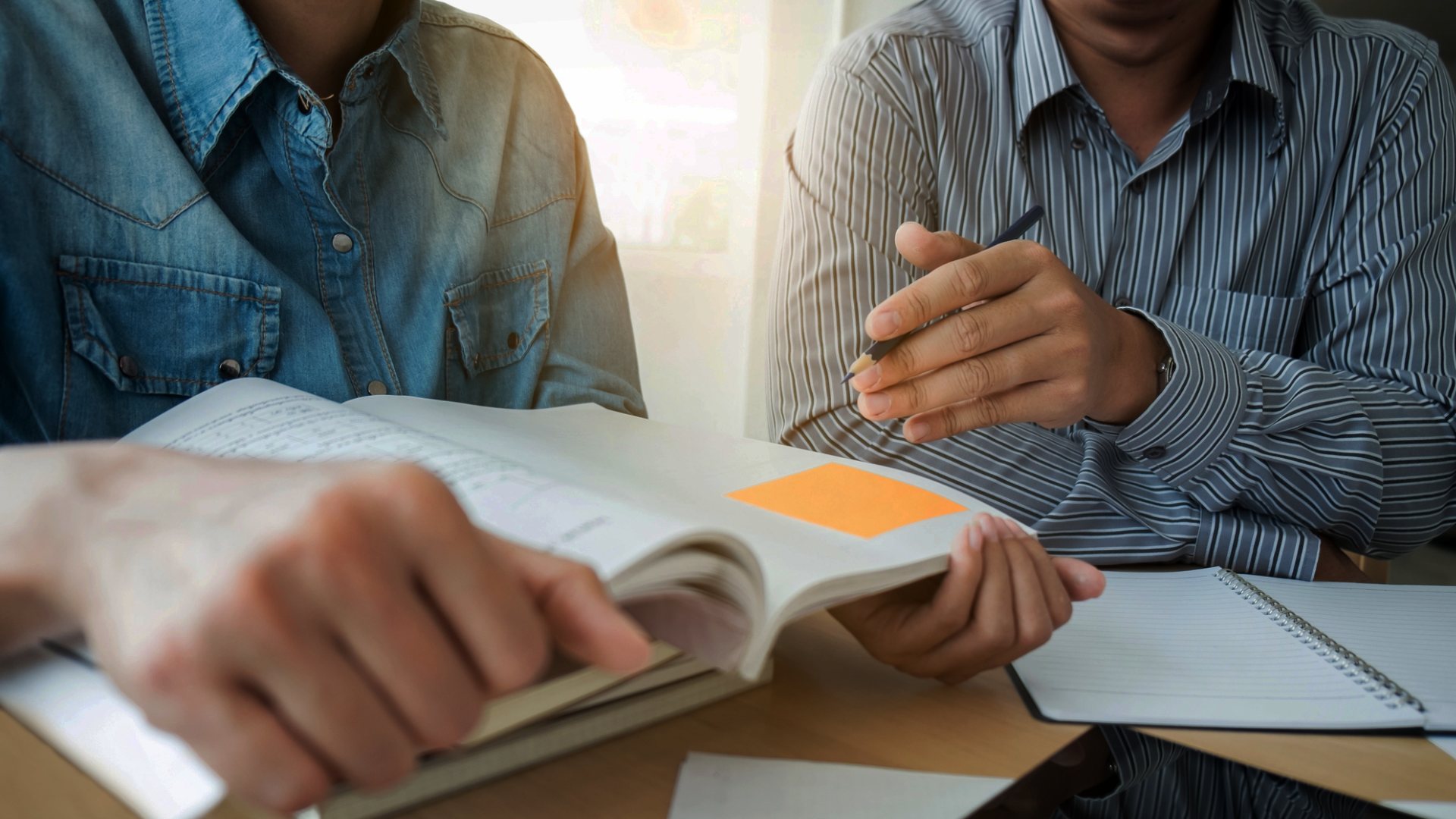 Två personer sitter vid bord och tittar i en bok med post it-lapp på.