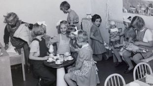 Svart-vit bild på flickor som leker med dockor i ett rum. Några tittar mot kameran och ler.