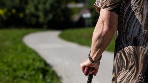 Äldre person går utomhus med käpp, klänning med bar arm, gräs på marken.