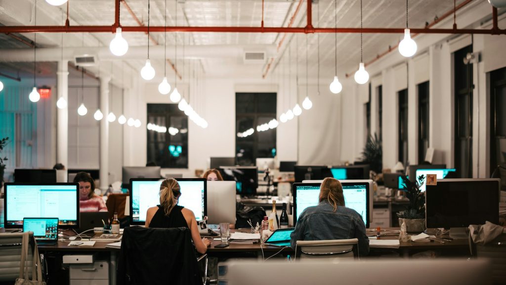 Öppet kontorslandskap med datorer och medarbetare, högt i tak.