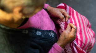 En gammal person sitter och virkar något randigt i utomhusmiljö, varmt solsken.