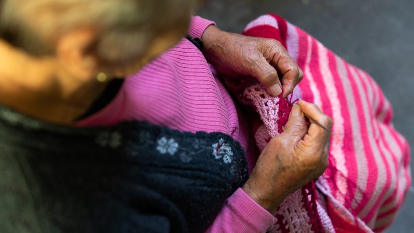 En gammal person sitter och virkar något randigt i utomhusmiljö, varmt solsken.